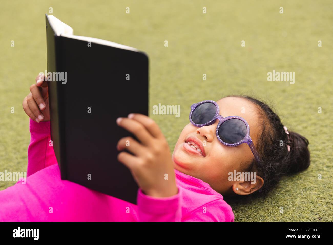 À l'école, jeune fille biraciale portant des lunettes de soleil couchées sur l'herbe à l'extérieur lisant Banque D'Images