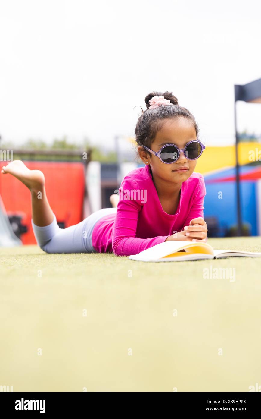 À l'école, une jeune fille biraciale portant des lunettes de soleil lit un livre sur l'herbe à l'extérieur Banque D'Images