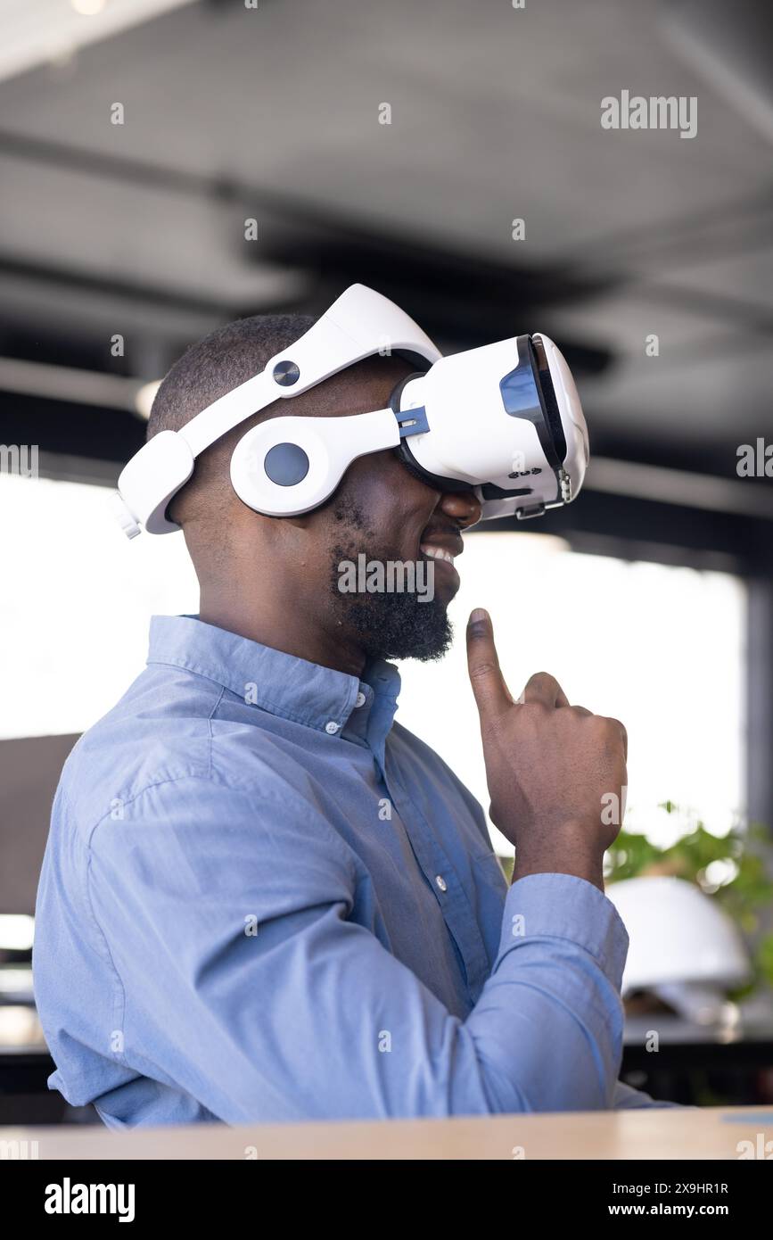 Jeune homme afro-américain portant casque de réalité virtuelle dans le bureau d'affaires. Il a les cheveux noirs courts, la barbe, et sourit en touchant le casque Banque D'Images