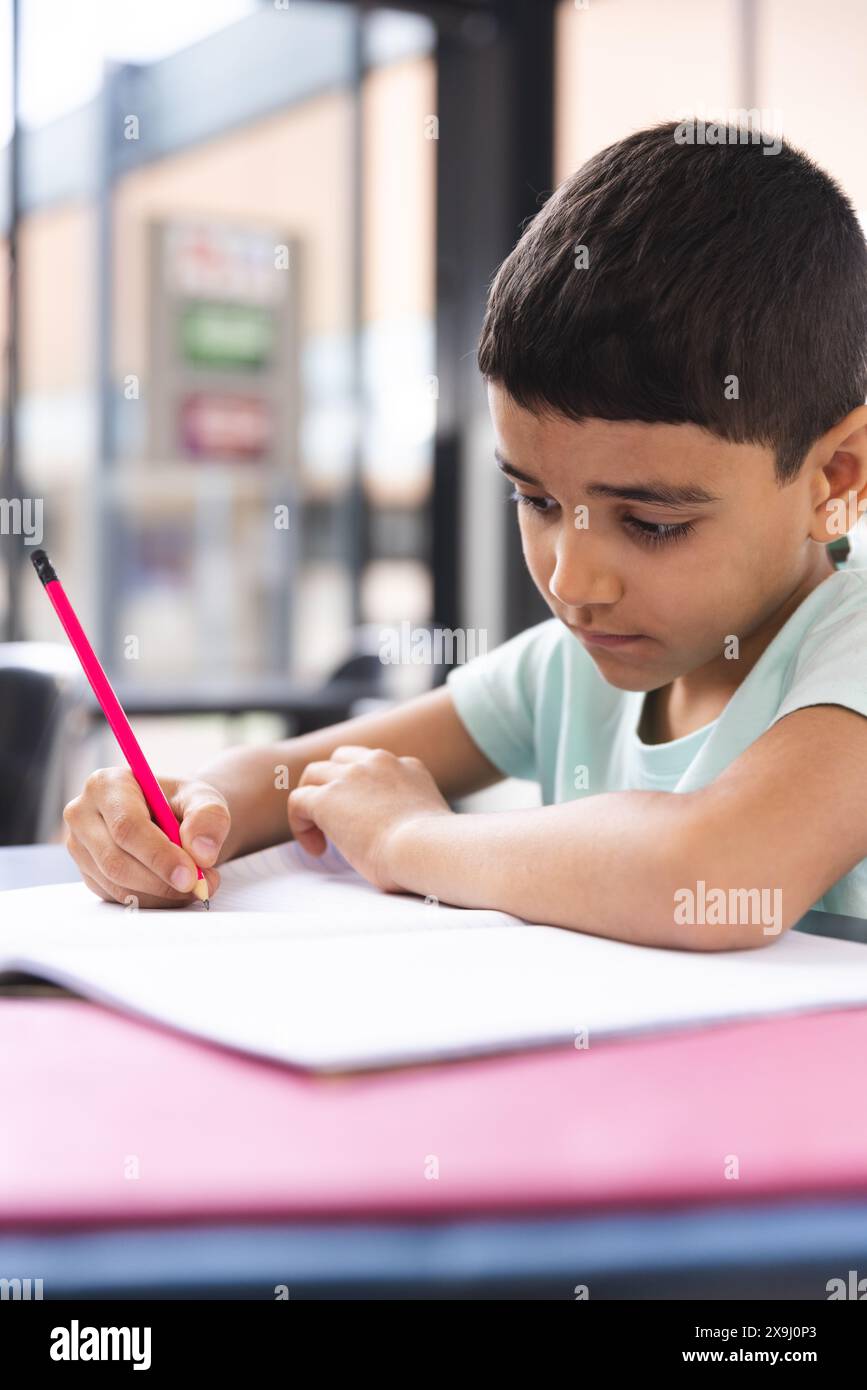 À l'école, jeune garçon biracial se concentrant sur l'écriture dans un cahier dans la salle de classe Banque D'Images