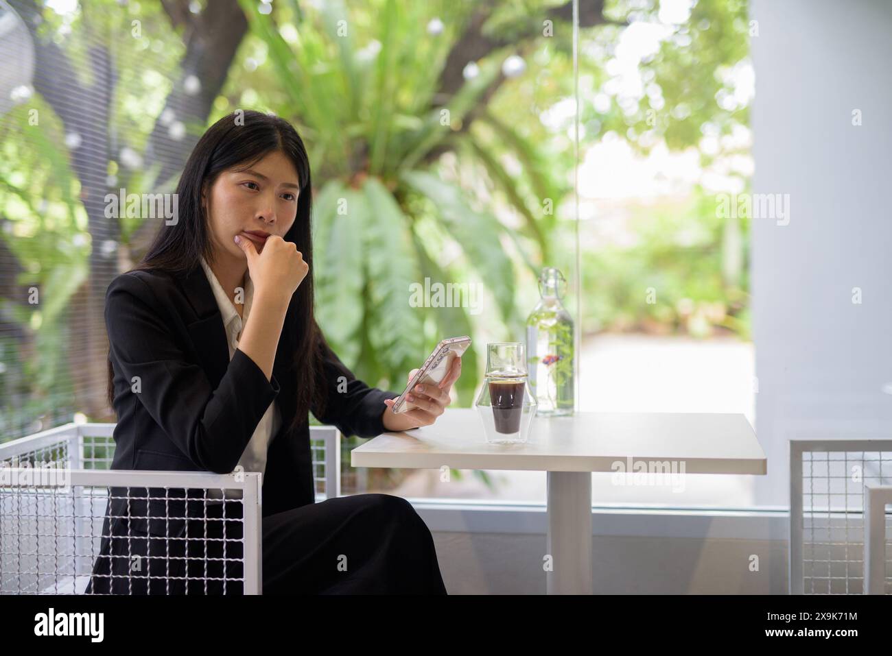 Femme d'affaires asiatique professionnelle travaillant sur son smartphone dans un café lumineux et moderne avec café et verdure Banque D'Images