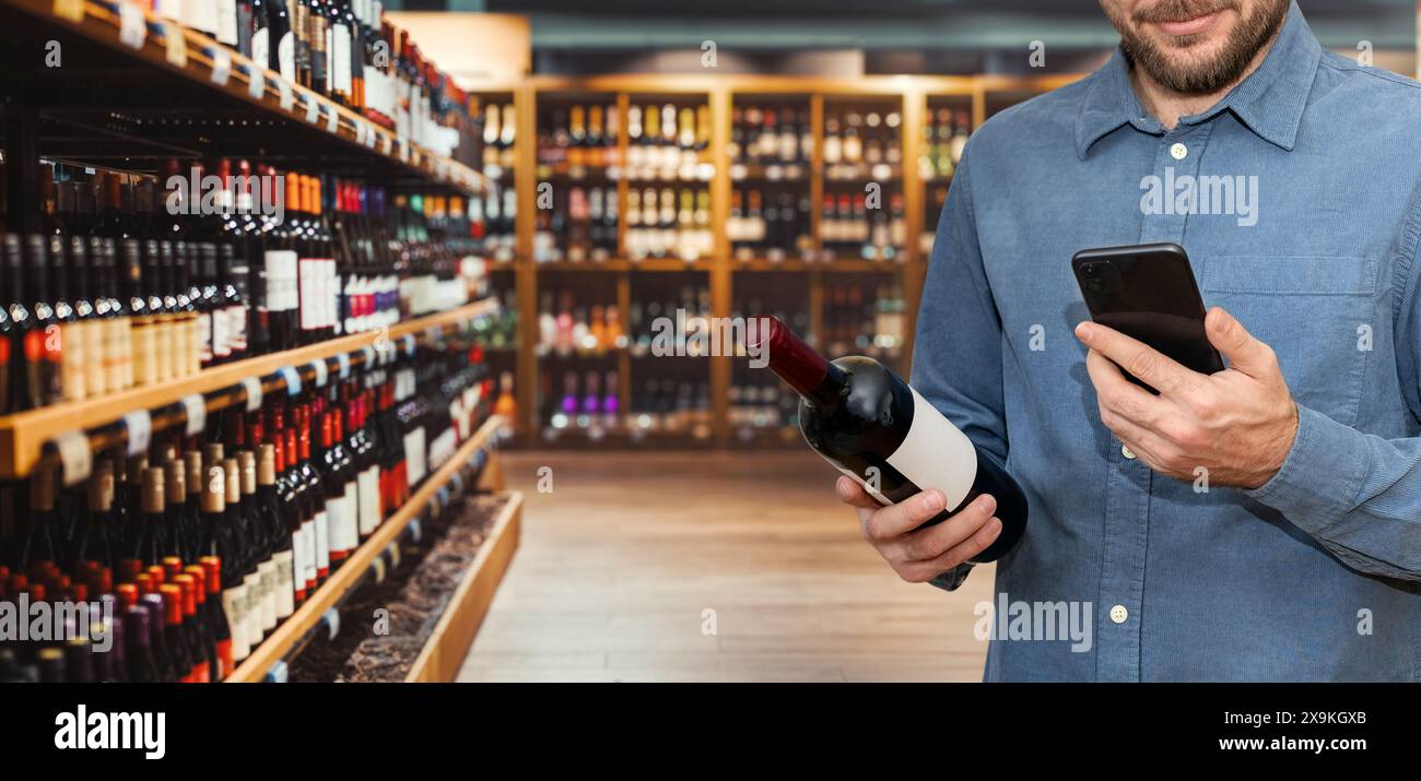 Client masculin achetant du vin dans un magasin d'alcool, tenant une bouteille de vin et un téléphone portable dans ses mains. Banque D'Images