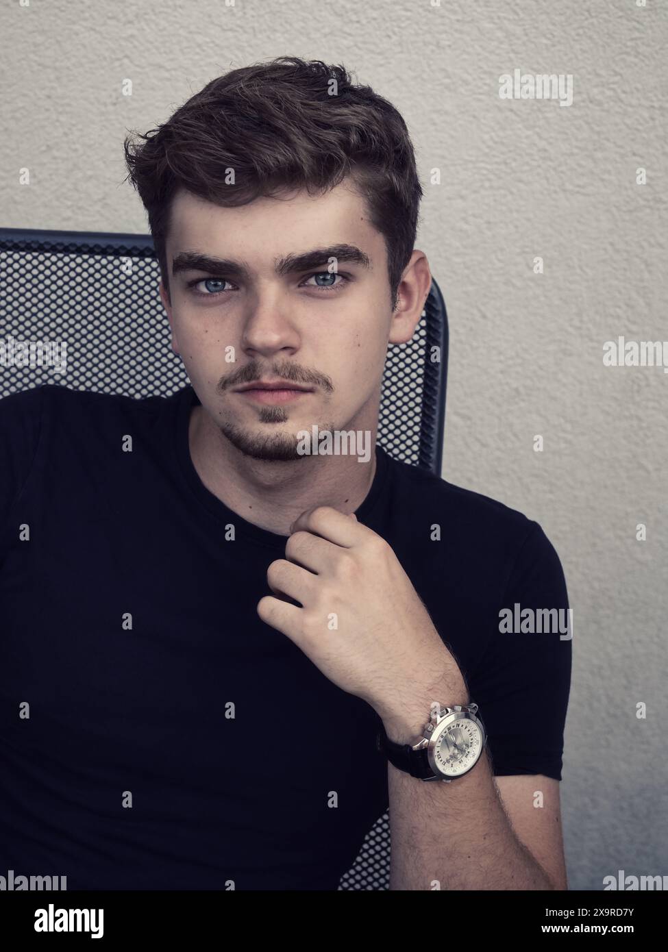 Portrait d'un jeune homme assis dehors contre un mur neutre, portant une chemise noire et une montre-bracelet. Image de style de vie décontracté avec expressio confiant Banque D'Images
