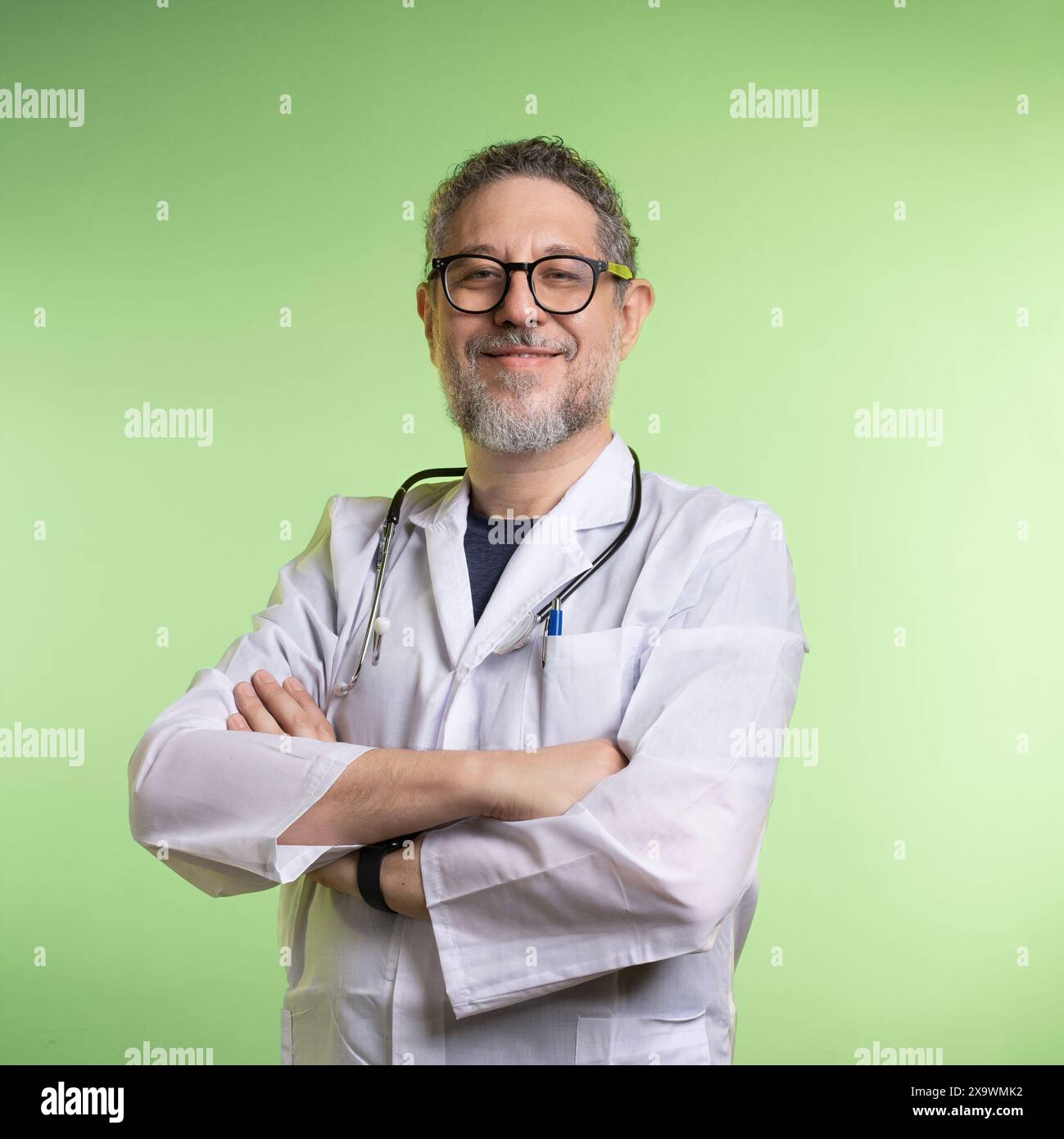 Médecin masculin souriant avec une barbe et des lunettes, portant une blouse de laboratoire et un stéthoscope, debout avec confiance avec les bras croisés, fond vert clair, repr Banque D'Images