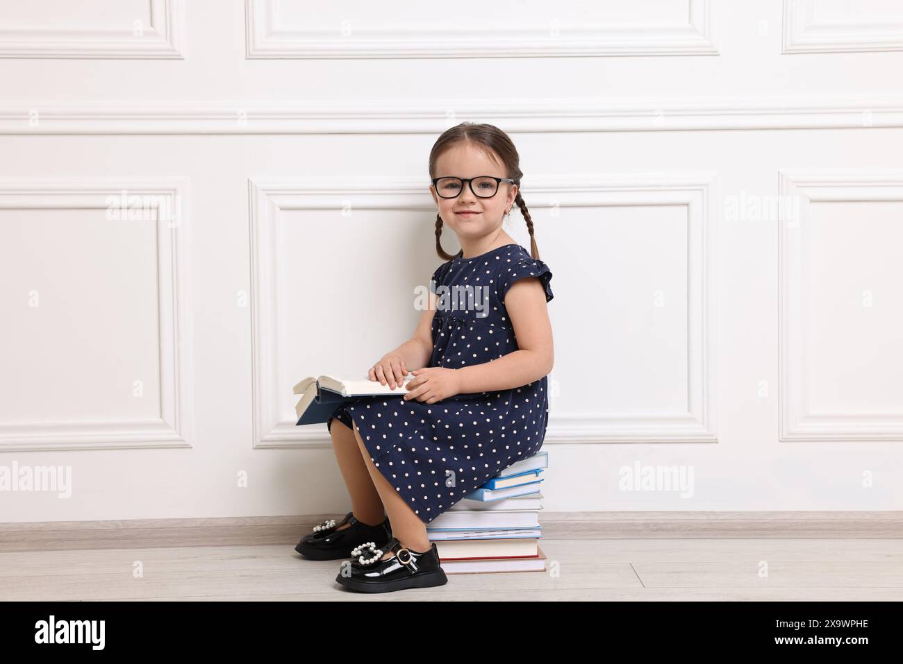 Mignonne petite fille dans des verres assis sur une pile de livres près du mur blanc Banque D'Images