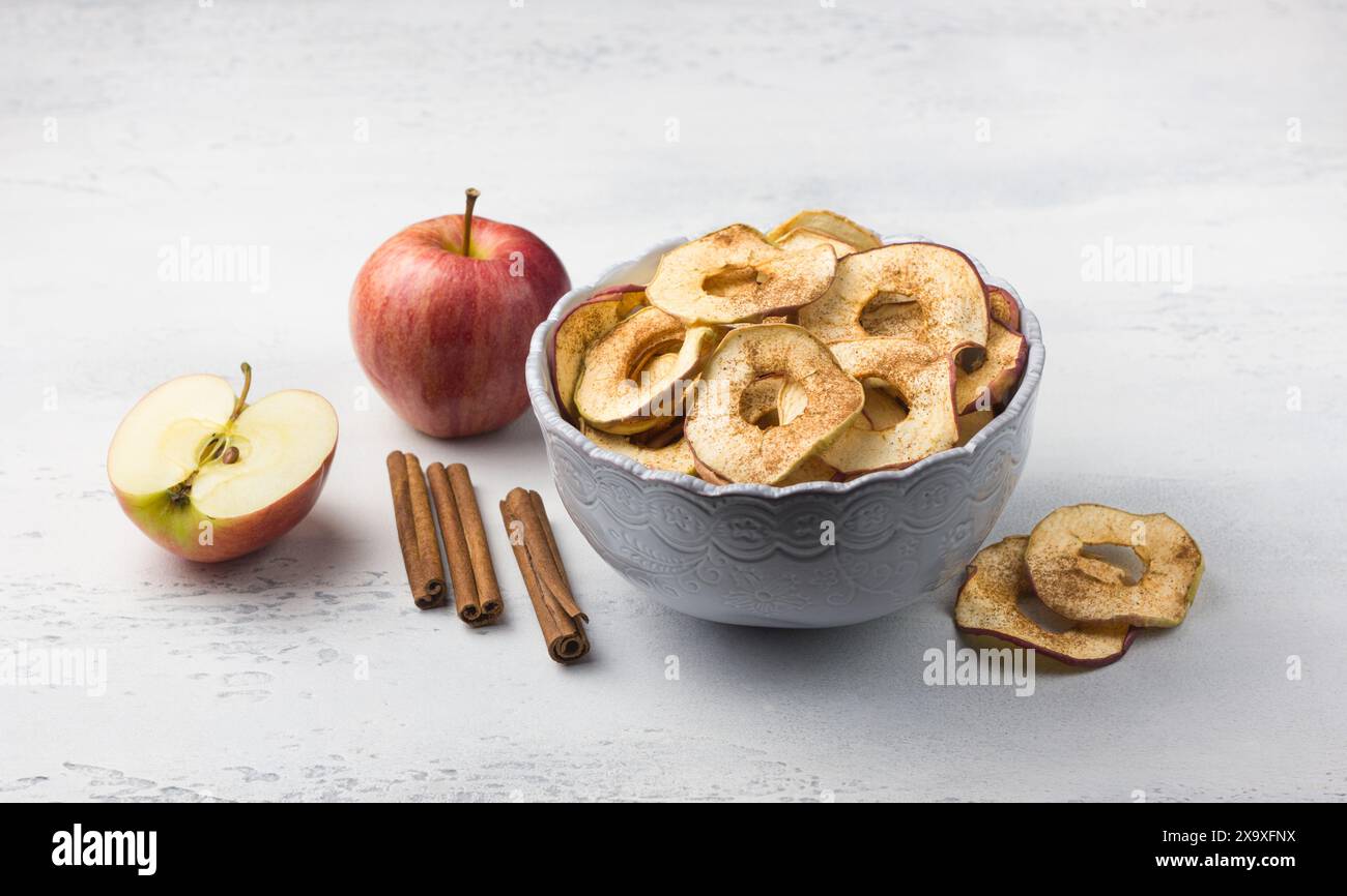 Pommes séchées maison à la cannelle, chips de pomme dans un bol avec des bâtons de cannelle sur un fond gris clair. Délicieux en-cas sain Banque D'Images
