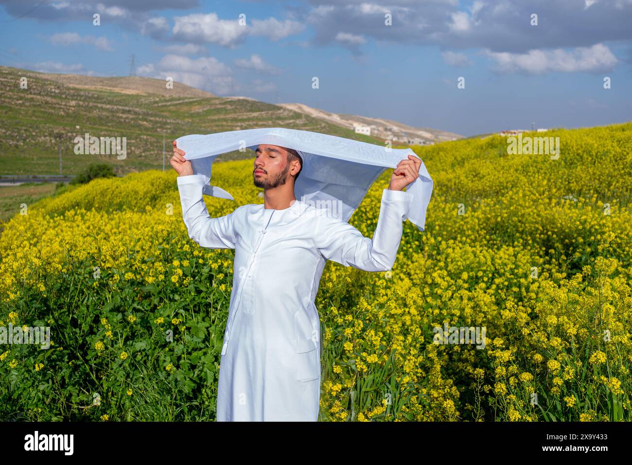 Un homme dans une dishdasha blanche et kandura se tient dans un champ de fleurs jaunes, soulevant la kandora les yeux fermés, profitant du soleil printanier, surrou Banque D'Images