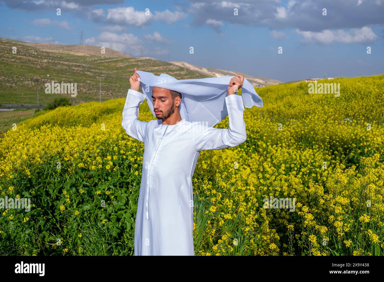 Un homme dans une dishdasha blanche et kandura se tient dans un champ de fleurs jaunes, soulevant la kandora les yeux fermés, profitant du soleil printanier, surrou Banque D'Images