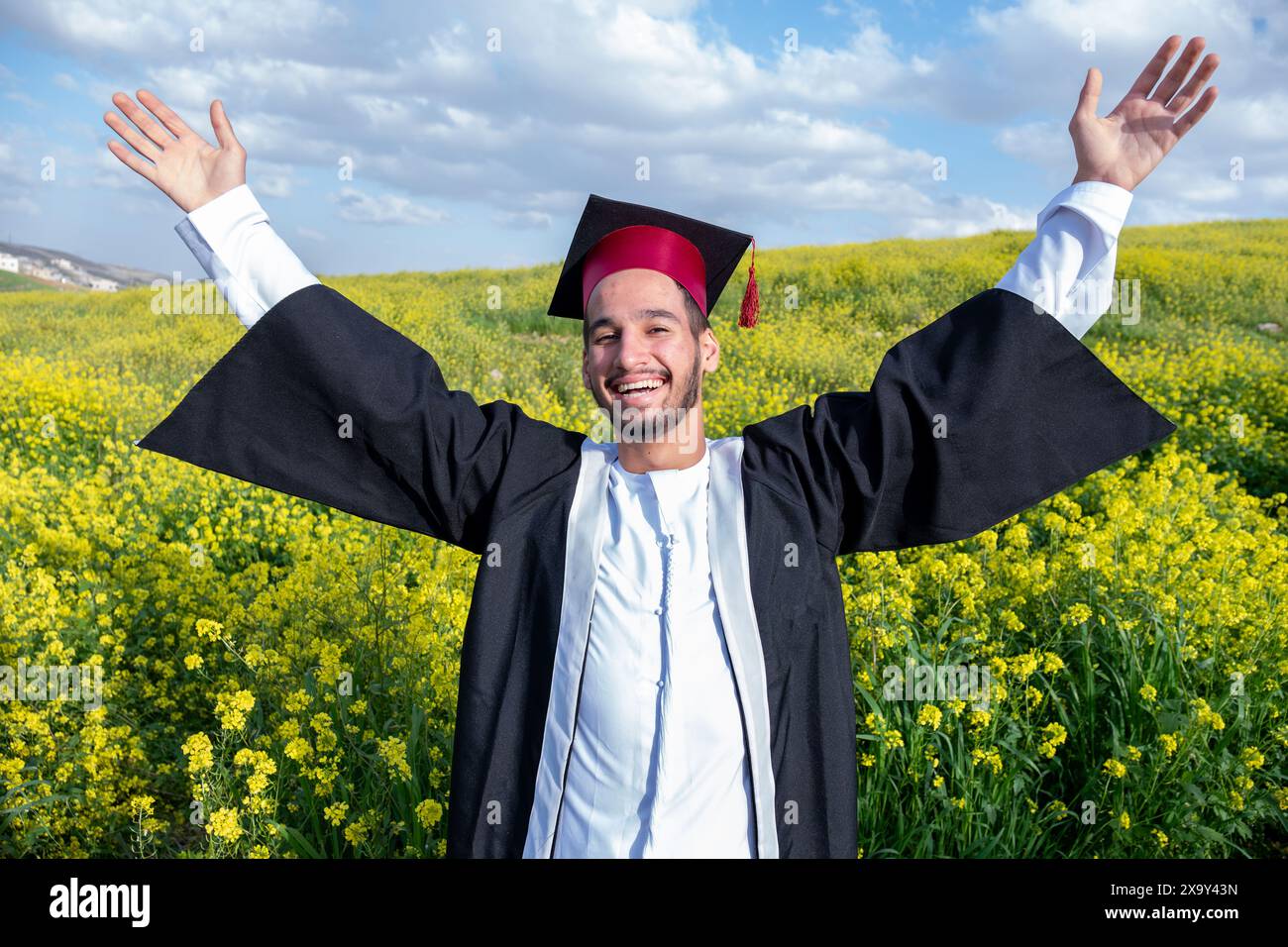 Joyeux jeune homme arabe portant kandora et célébrant son diplôme dans un beau champ de roses jaunes avec le sourire sur son visage levant son poing à th Banque D'Images