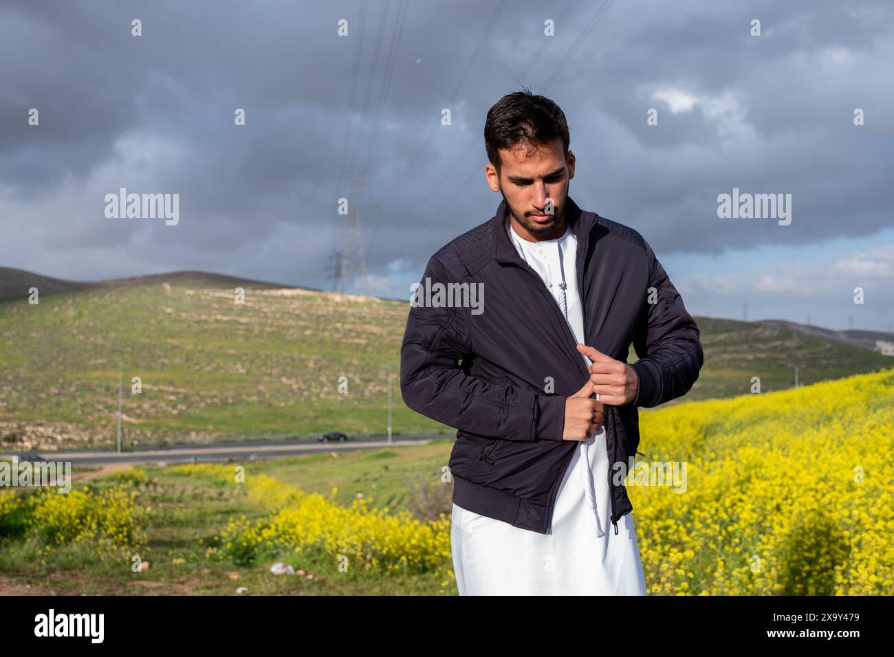 Un homme dans une veste noire et dishdasha se tient dans un champ vibrant de fleurs jaunes et de collines vertes, les bras croisés pour la chaleur, sous un ciel de printemps nuageux, Banque D'Images