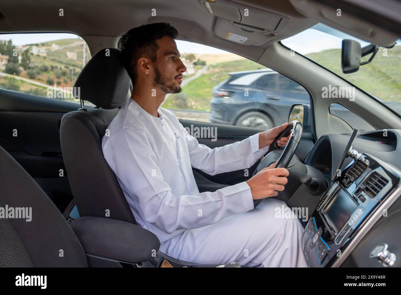 Un jeune homme joyeux avec une barbe, portant une tenue traditionnelle du moyen-Orient, sourit en toute confiance en étant assis dans sa voiture. Il semble s'amuser Banque D'Images