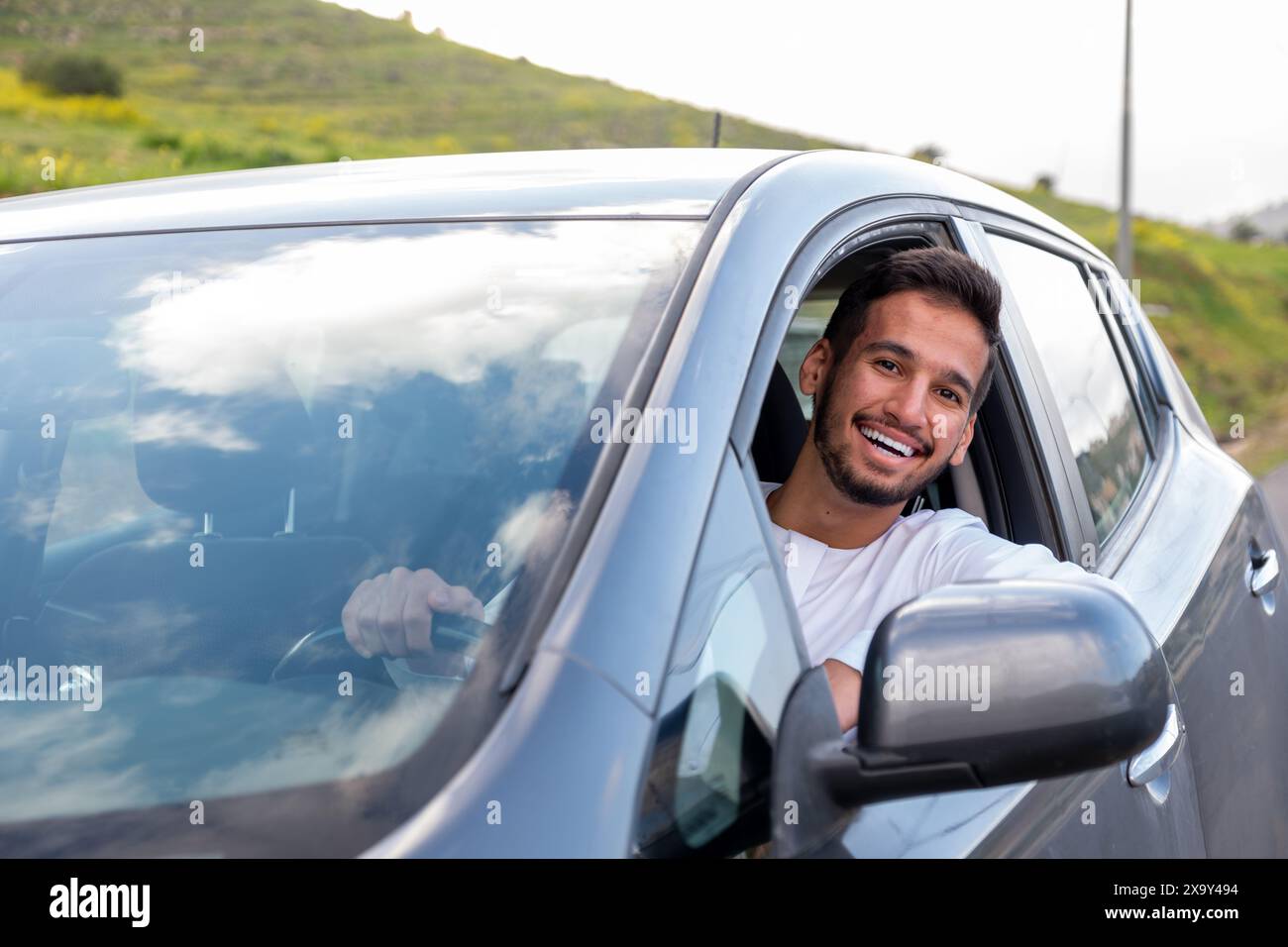 Un jeune homme joyeux avec une barbe, portant une tenue traditionnelle du moyen-Orient, sourit en toute confiance en étant assis dans sa voiture. Il semble s'amuser Banque D'Images