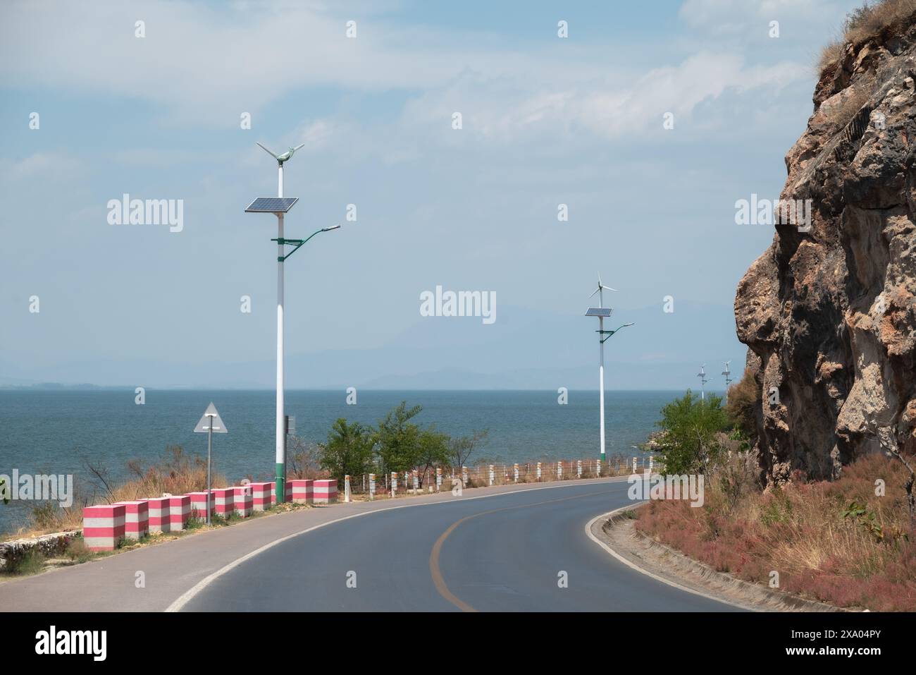 Une route courbe près de la mer avec de petites éoliennes et des panneaux solaires sur les poteaux Banque D'Images