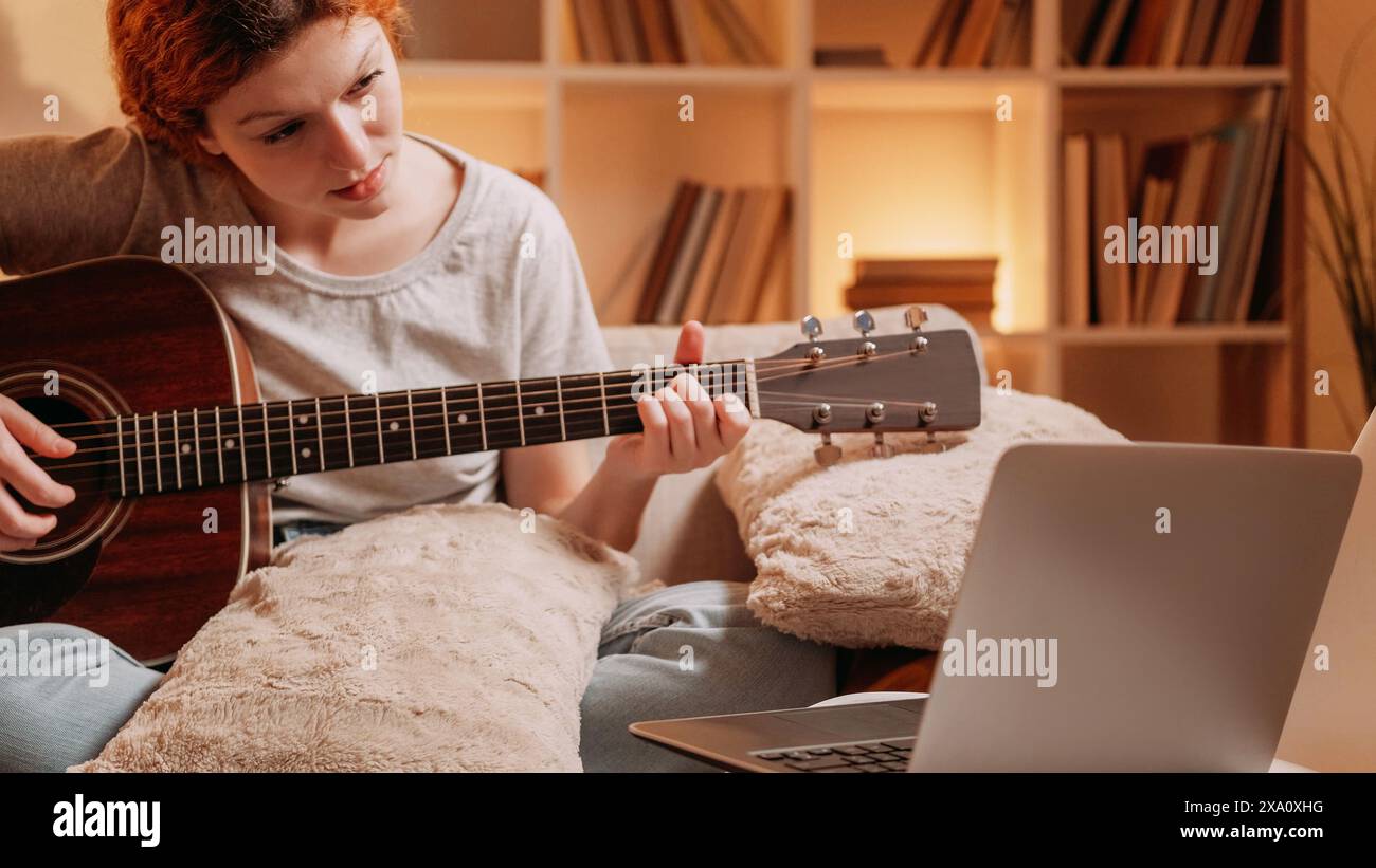 Cours de musique en ligne. Cours acoustique. Passe-temps créatif. Femme musicienne inspirée jouant des accords d'apprentissage de guitare regardant la leçon virtuelle sur ordinateur portable sur canapé à la maison. Banque D'Images