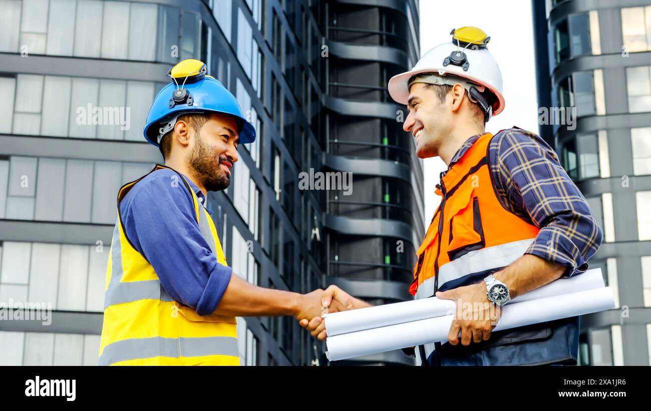 Les deux ouvriers de la construction en casques de sécurité se serrent la main à l'extérieur avec des plans. Banque D'Images