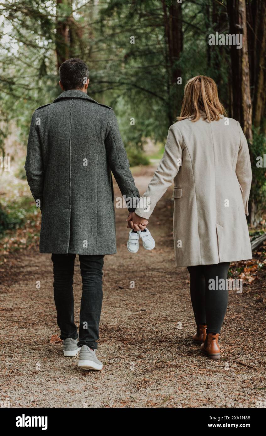 Séance photo de maternité avec couple se tenant la main et des baskets de bébé blanches tout en marchant dans le parc. Banque D'Images