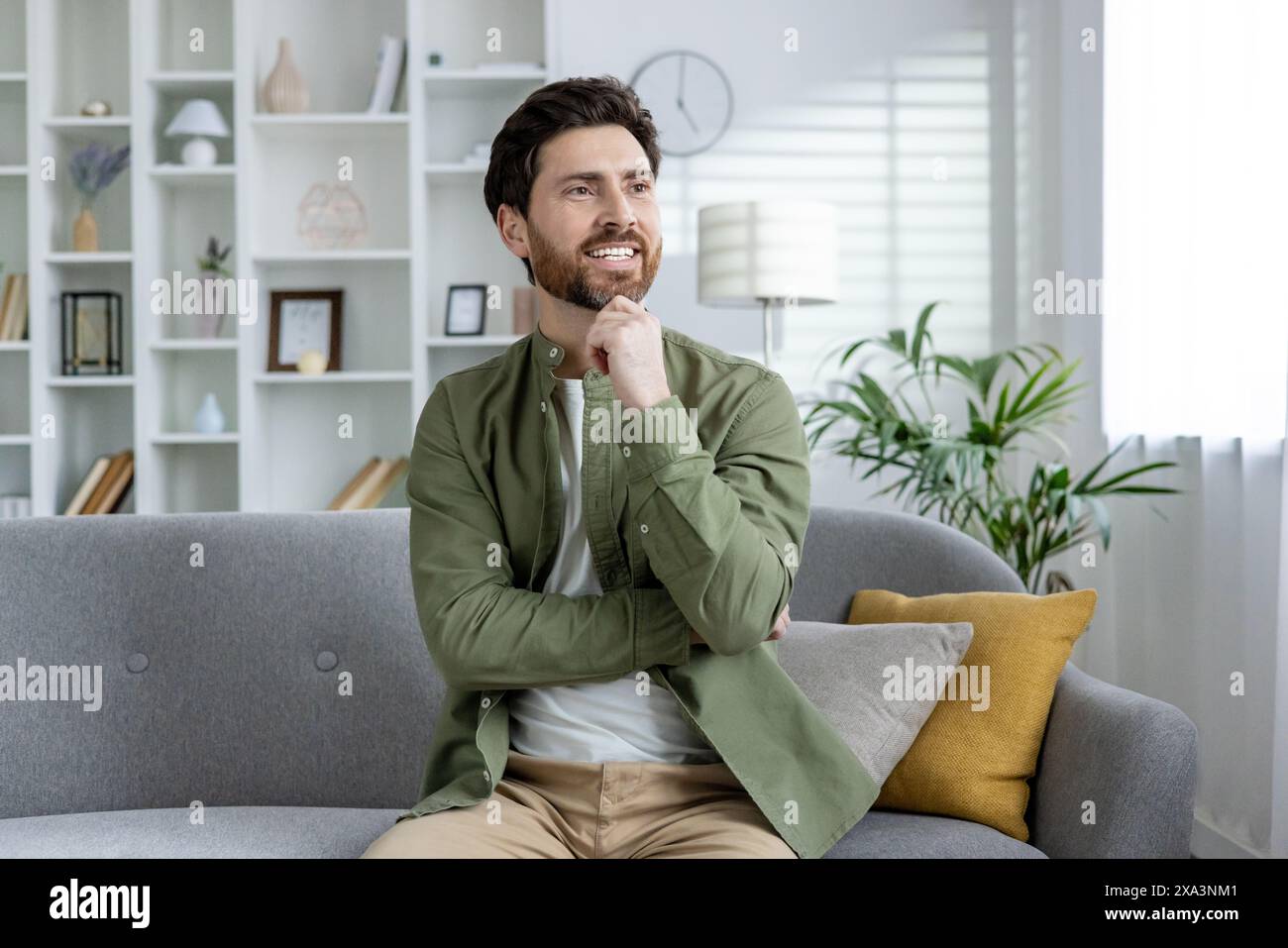 Homme barbu assis sur un canapé gris dans un salon élégant, perdu dans la pensée et le sourire. Intérieur de maison lumineux et confortable avec des plantes et un décor. Banque D'Images