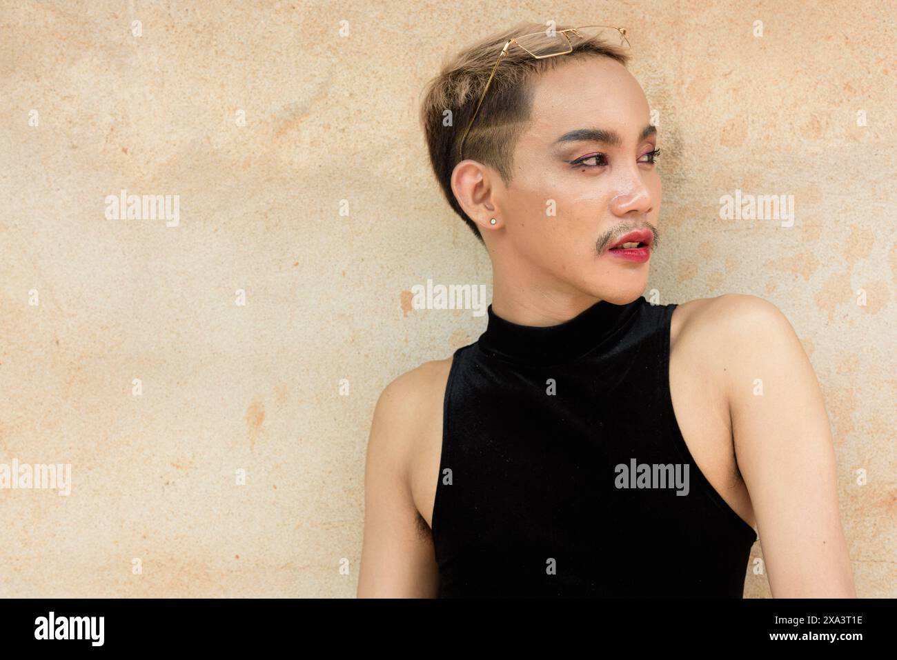 Portrait d'un bel homme asiatique de soutien de la communauté LGBT queer avec moustache portant rouge à lèvres et combinaison sur fond de mur beige Banque D'Images