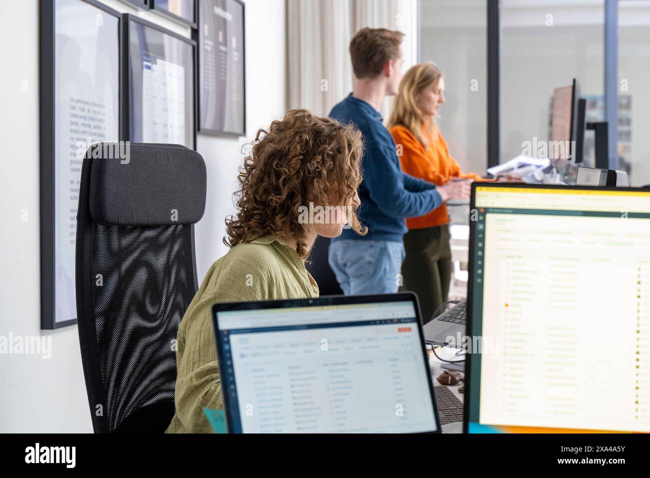 Trois professionnels travaillant dans un espace de bureau moderne, l'un se concentrant sur un écran d'ordinateur au premier plan et deux collègues discutant d'un document en arrière-plan. Banque D'Images