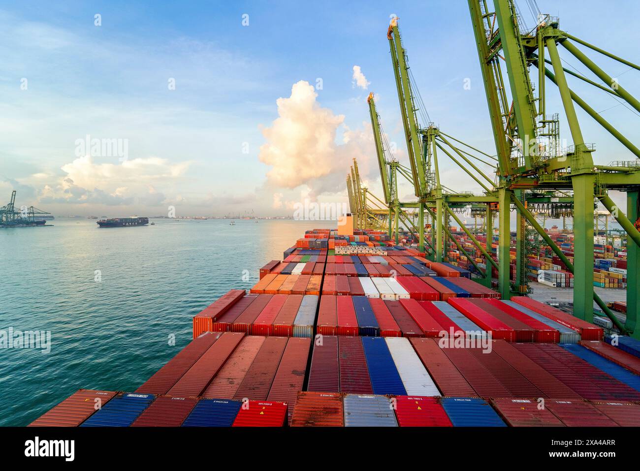 Conteneurs de fret empilés sur un grand navire dans un port au lever du soleil, avec des grues et un ciel dégagé en arrière-plan. Banque D'Images