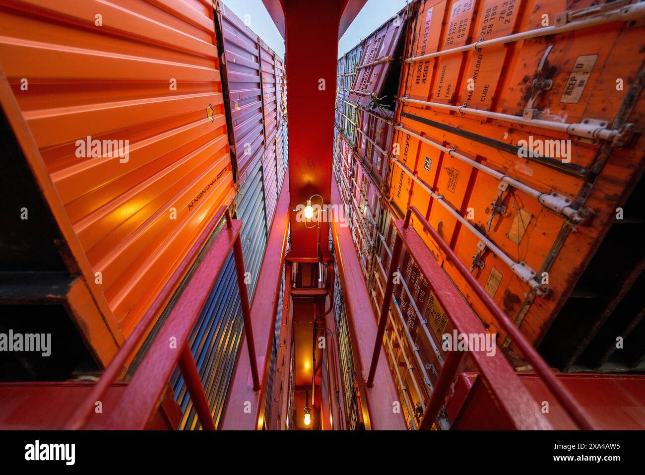 Une vue depuis le bas d'une allée étroite encadrée par des piles imposantes de conteneurs d'expédition colorés au crépuscule, avec un lampadaire éclairé au centre. Banque D'Images
