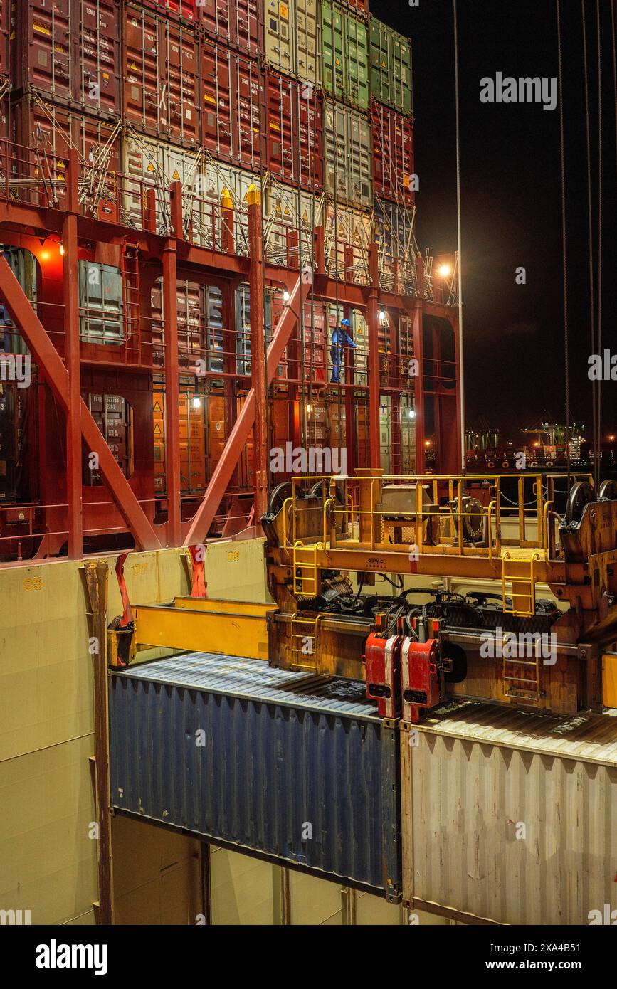 Une vue nocturne d'un port à conteneurs animé, avec un navire en cours de chargement ou de déchargement par une grande grue à portique éclairée, remplie de conteneurs colorés. Banque D'Images