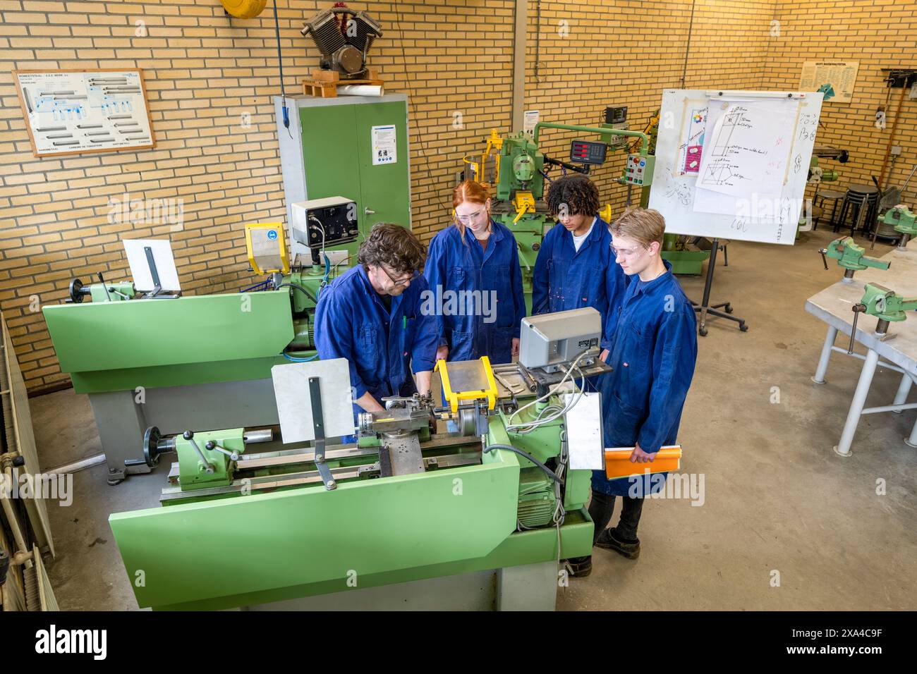 Quatre personnes en tenue de travail bleue utilisent des machines industrielles vertes dans un atelier, collaborant sur une tâche technique. Banque D'Images