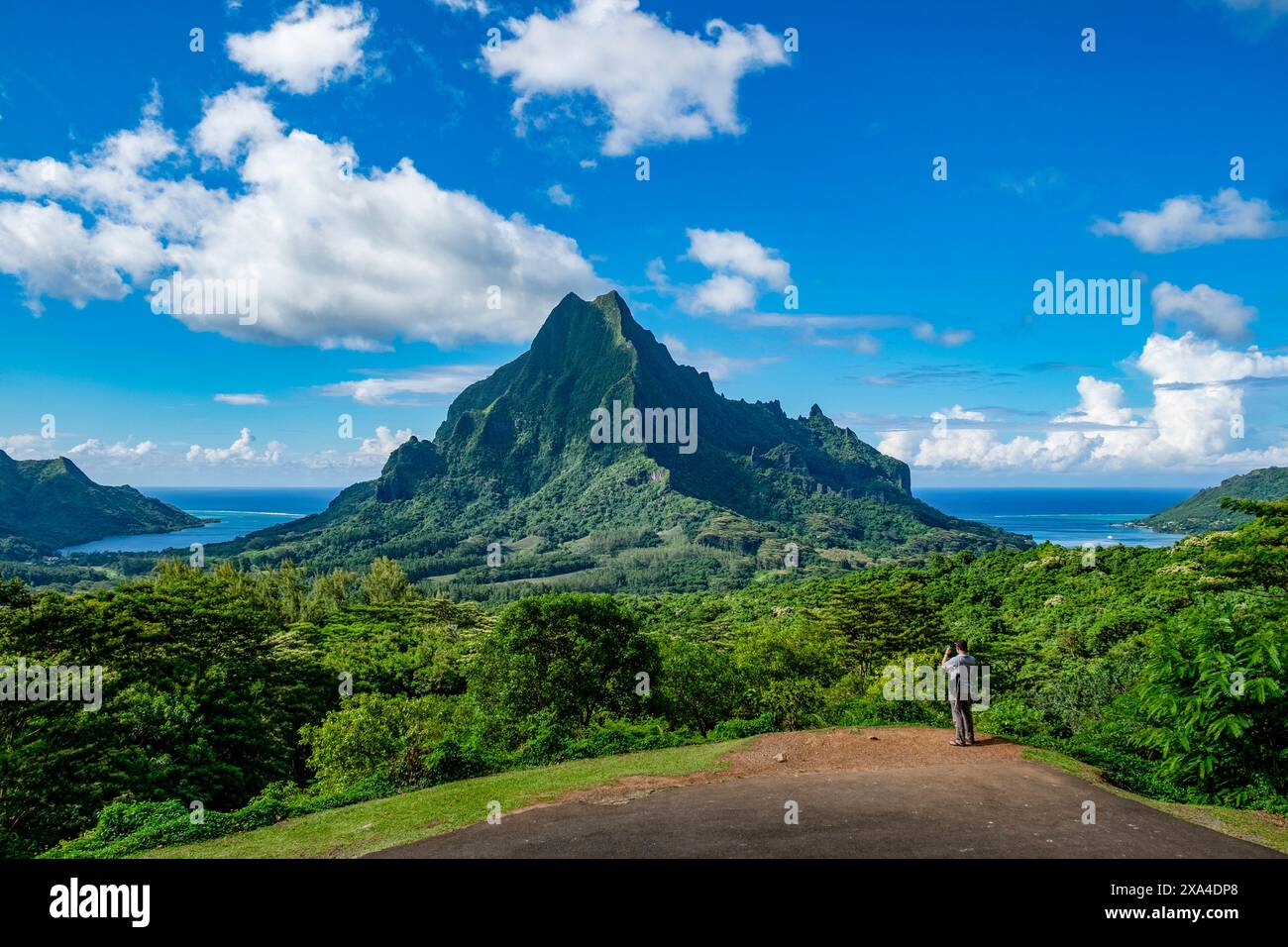 Une personne se tient sur un belvédère admirant la vue imprenable sur un sommet de montagne luxuriant et vert sous un ciel bleu vibrant avec quelques nuages. Banque D'Images