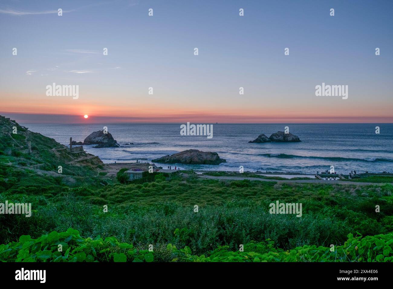 Vue sur le coucher du soleil sur un paysage côtier avec des collines ondulantes, une végétation luxuriante au premier plan, et plusieurs formations rocheuses dans l'océan. Banque D'Images