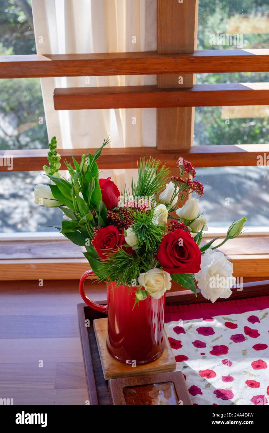 Un bouquet vibrant de fleurs avec des roses rouges et des tulipes blanches dans une tasse rouge, placé sur une surface en bois près d'une fenêtre avec des rideaux transparents et une vue sur la verdure à l'extérieur. Banque D'Images