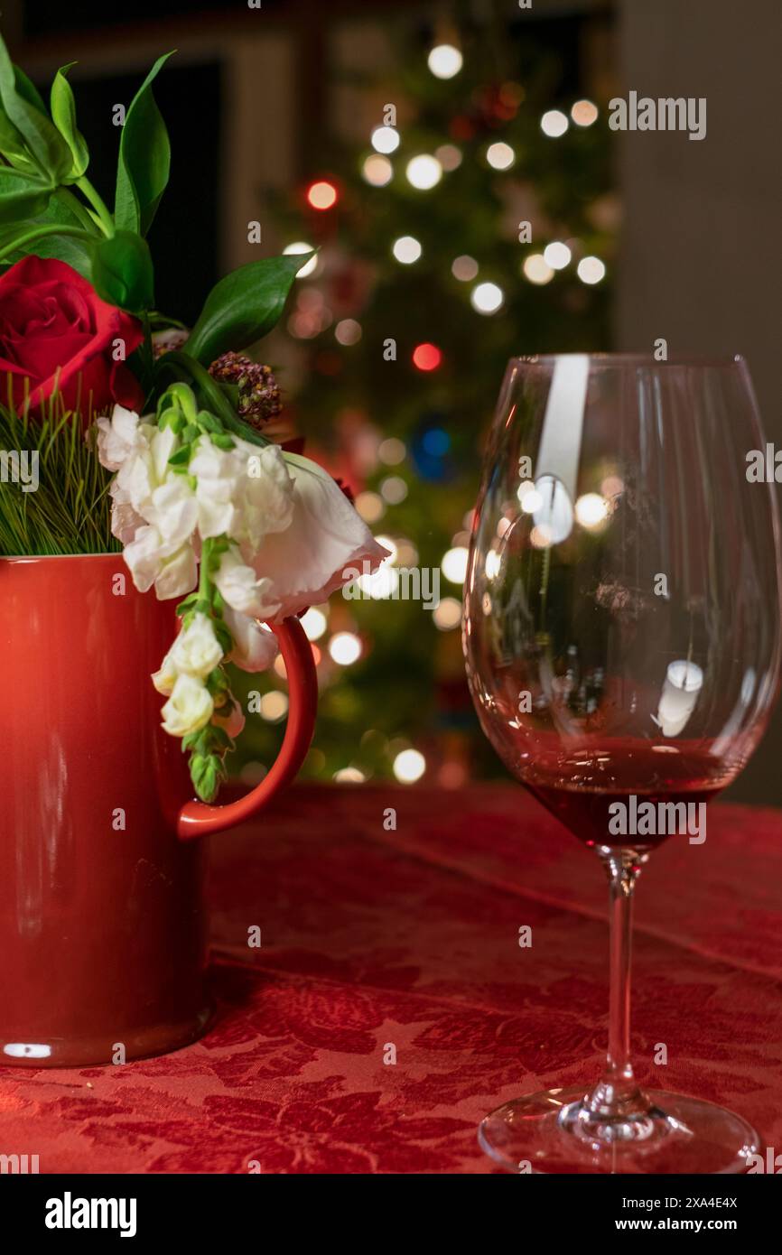 Une table festive avec un bouquet de fleurs dans un vase rouge et un verre à vin vide, avec un sapin de Noël flou orné de lumières en arrière-plan. Banque D'Images