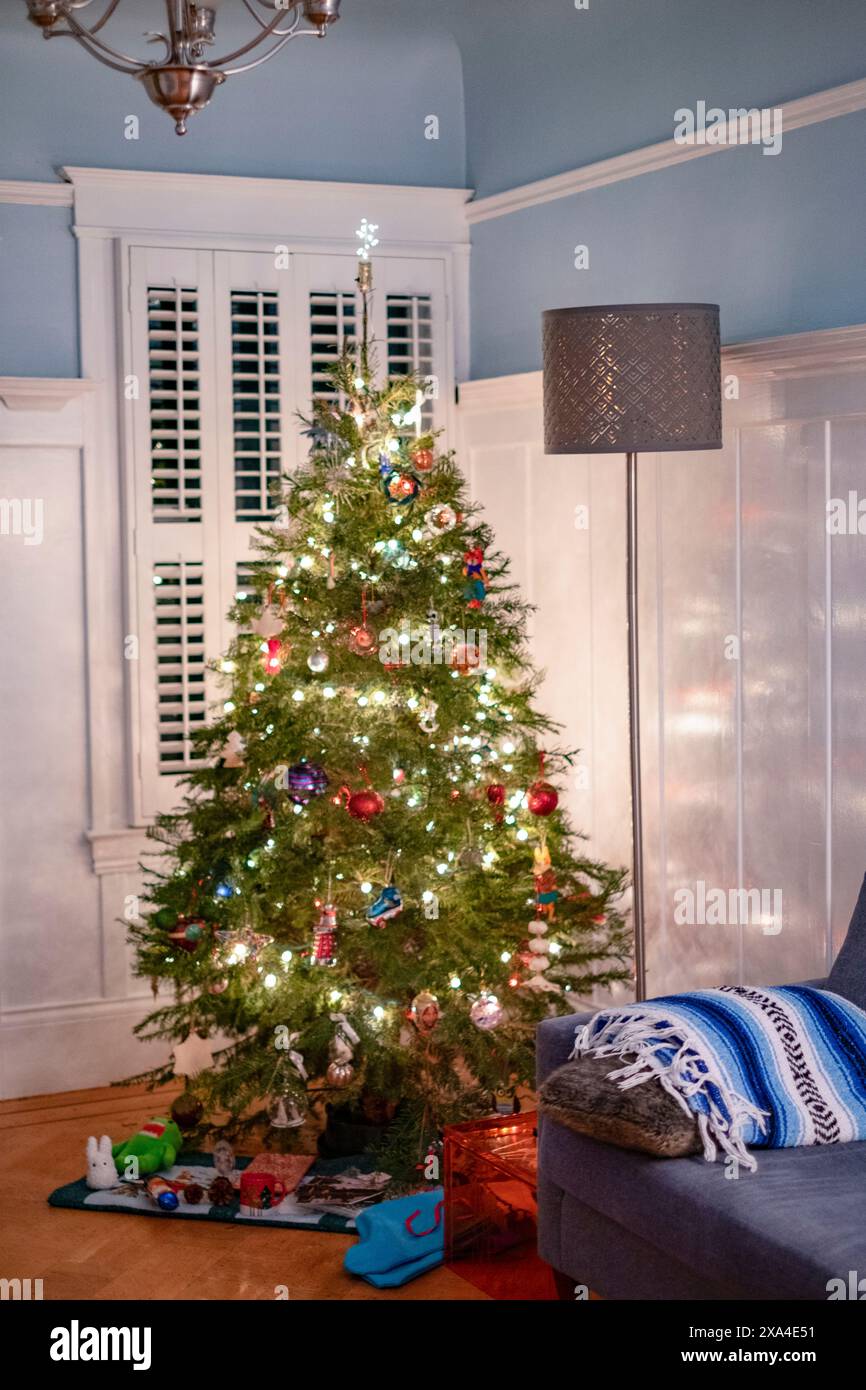 L'image présente un arbre de Noël décoré avec des ornements colorés et des lumières dans une pièce confortable avec un mur bleu clair, une fenêtre avec des volets blancs et un canapé gris avec une couverture bleue et blanche sur le côté. Banque D'Images