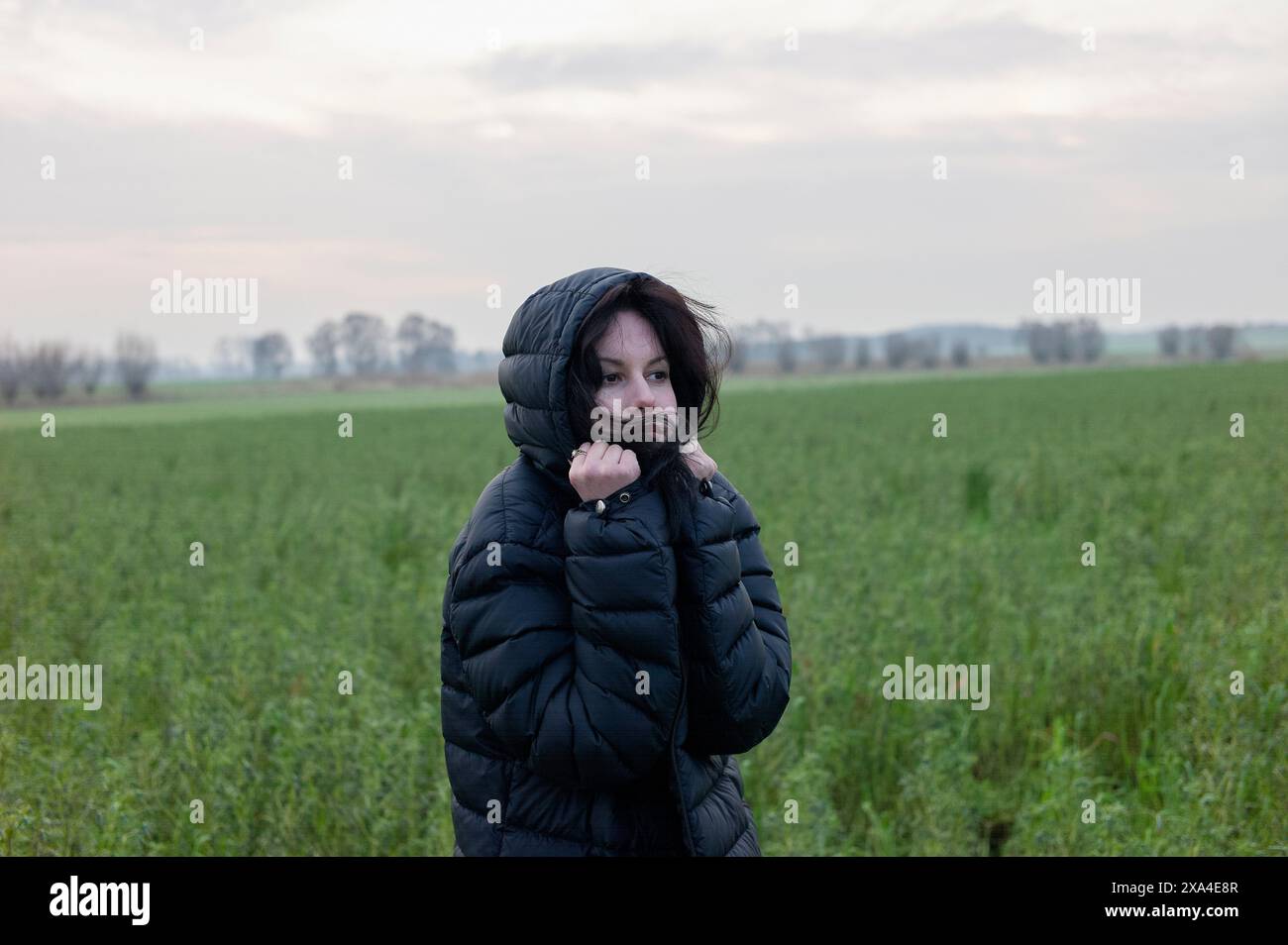 Une femme se tient dans un champ vert avec un ciel nuageux au-dessus de la tête, portant une veste bouffante noire et couvrant partiellement son visage avec son col. Banque D'Images
