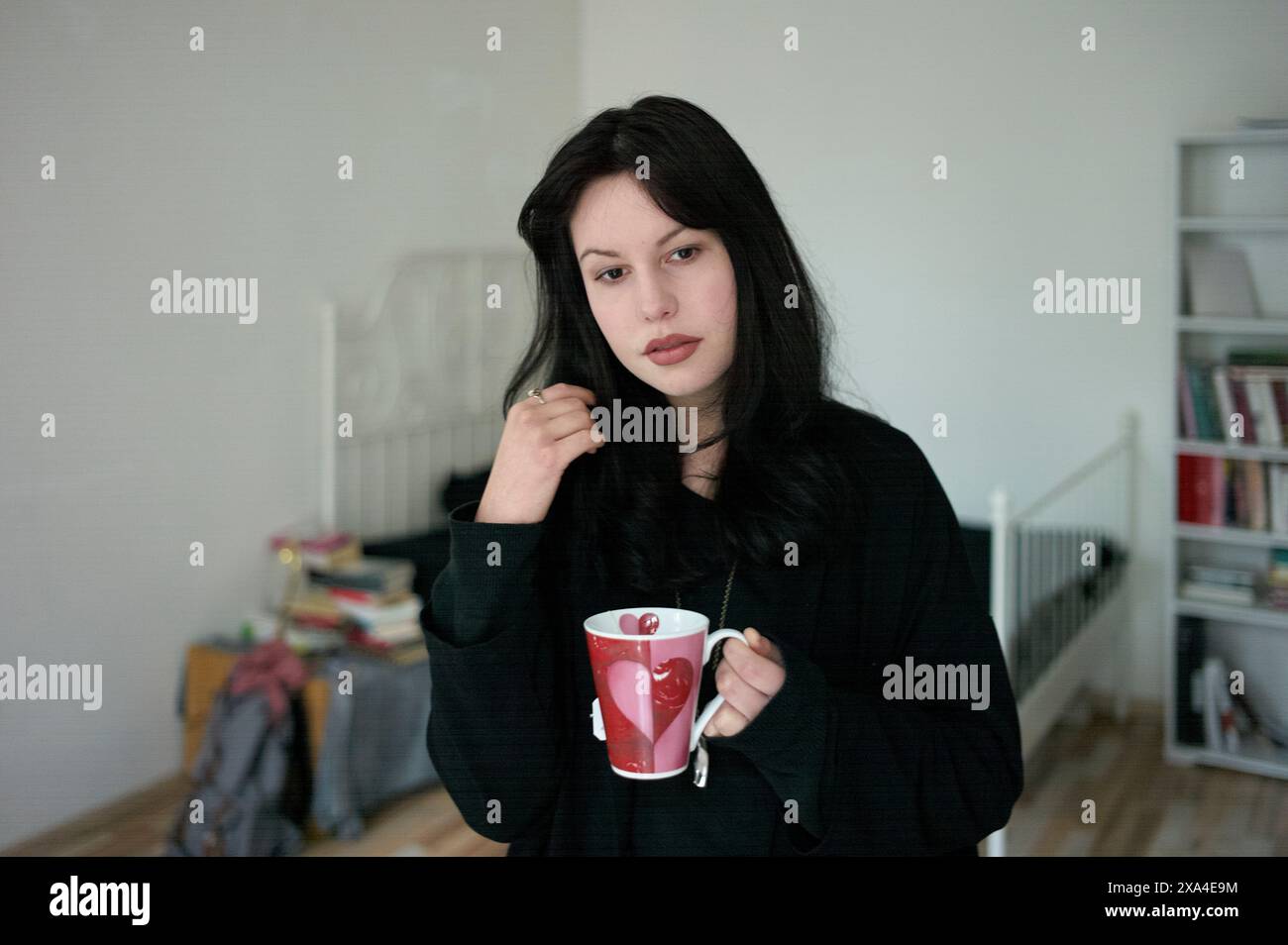 Une femme aux cheveux foncés tient une tasse rouge, debout dans une pièce avec une étagère blanche en arrière-plan. Banque D'Images