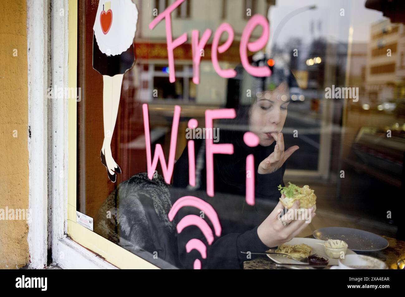 Une personne vue à travers une fenêtre s'assoit à l'intérieur d'un café, mangeant un sandwich avec un panneau WiFi gratuit peint sur le verre.' Banque D'Images