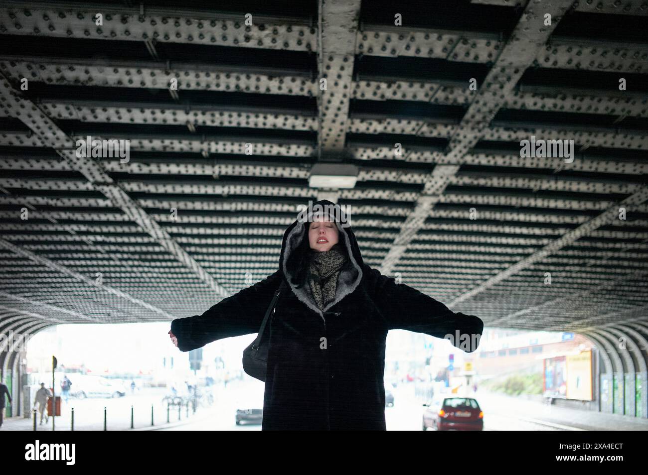 Une personne se tient debout avec les bras tendus sous un pont urbain avec un plafond à motifs, les yeux fermés, portant un manteau d'hiver et une écharpe. Banque D'Images