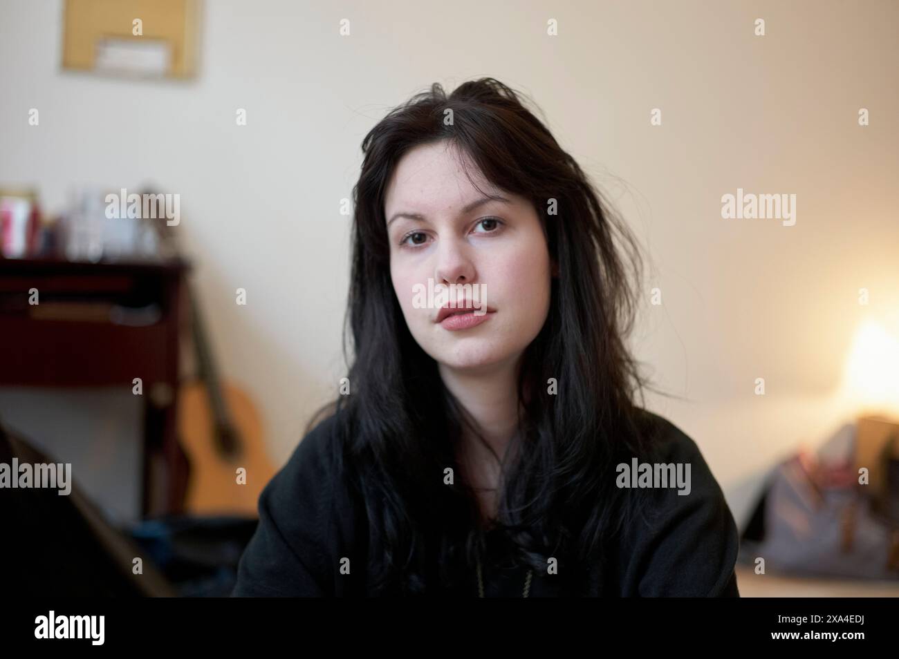 Une femme aux cheveux foncés est assise dans une pièce, avec certains éléments tels que des images et une guitare floue en arrière-plan. Banque D'Images