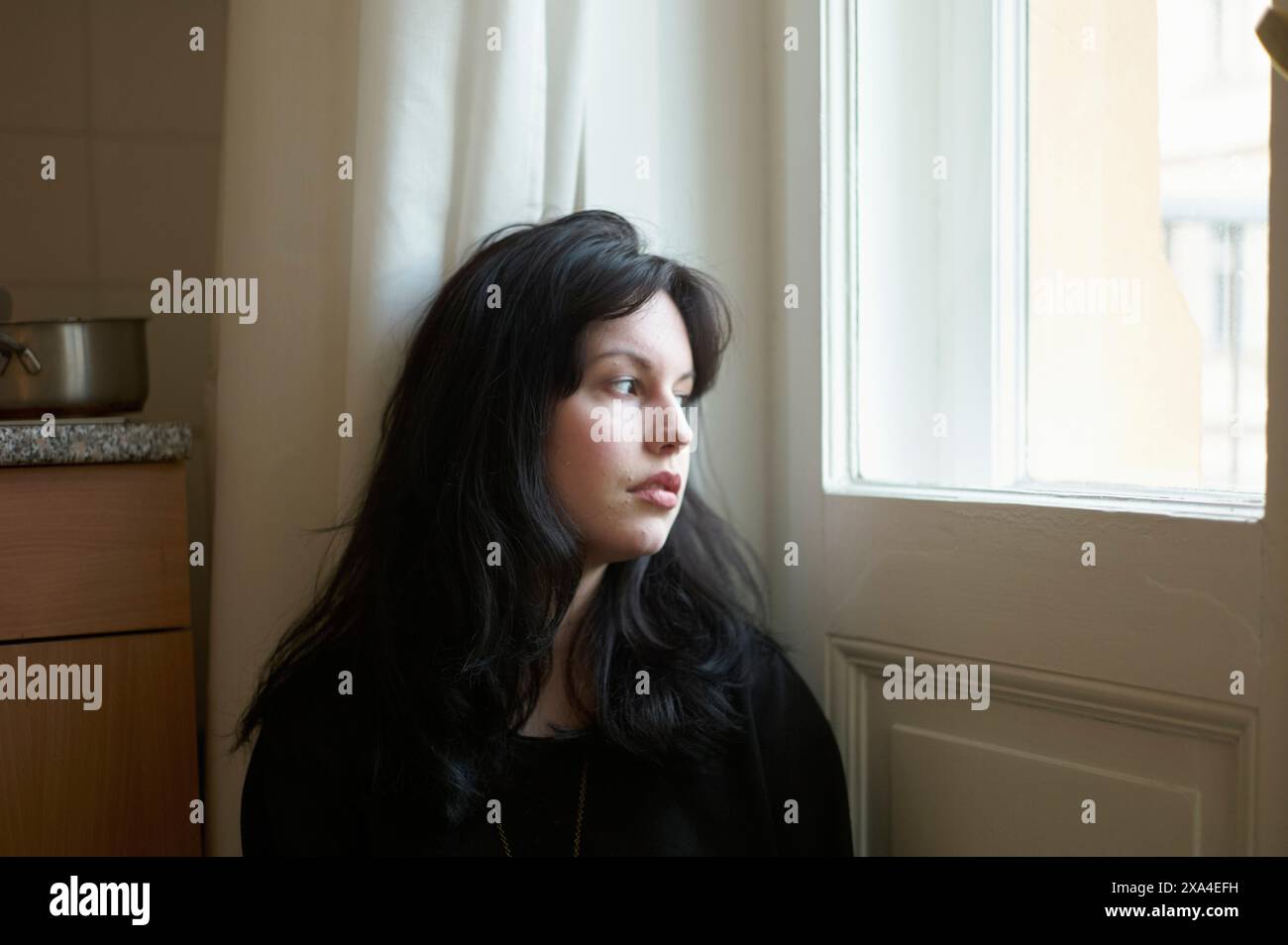 Une femme aux cheveux noirs regarde par une fenêtre, son visage éclairé par la lumière naturelle. Banque D'Images
