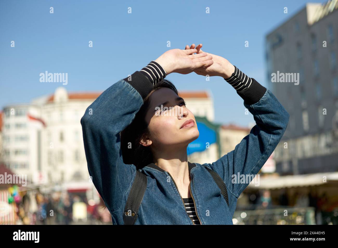 Une femme protège ses yeux du soleil avec sa main, regardant vers le haut contre une toile de fond de bâtiments et un ciel bleu clair. Banque D'Images