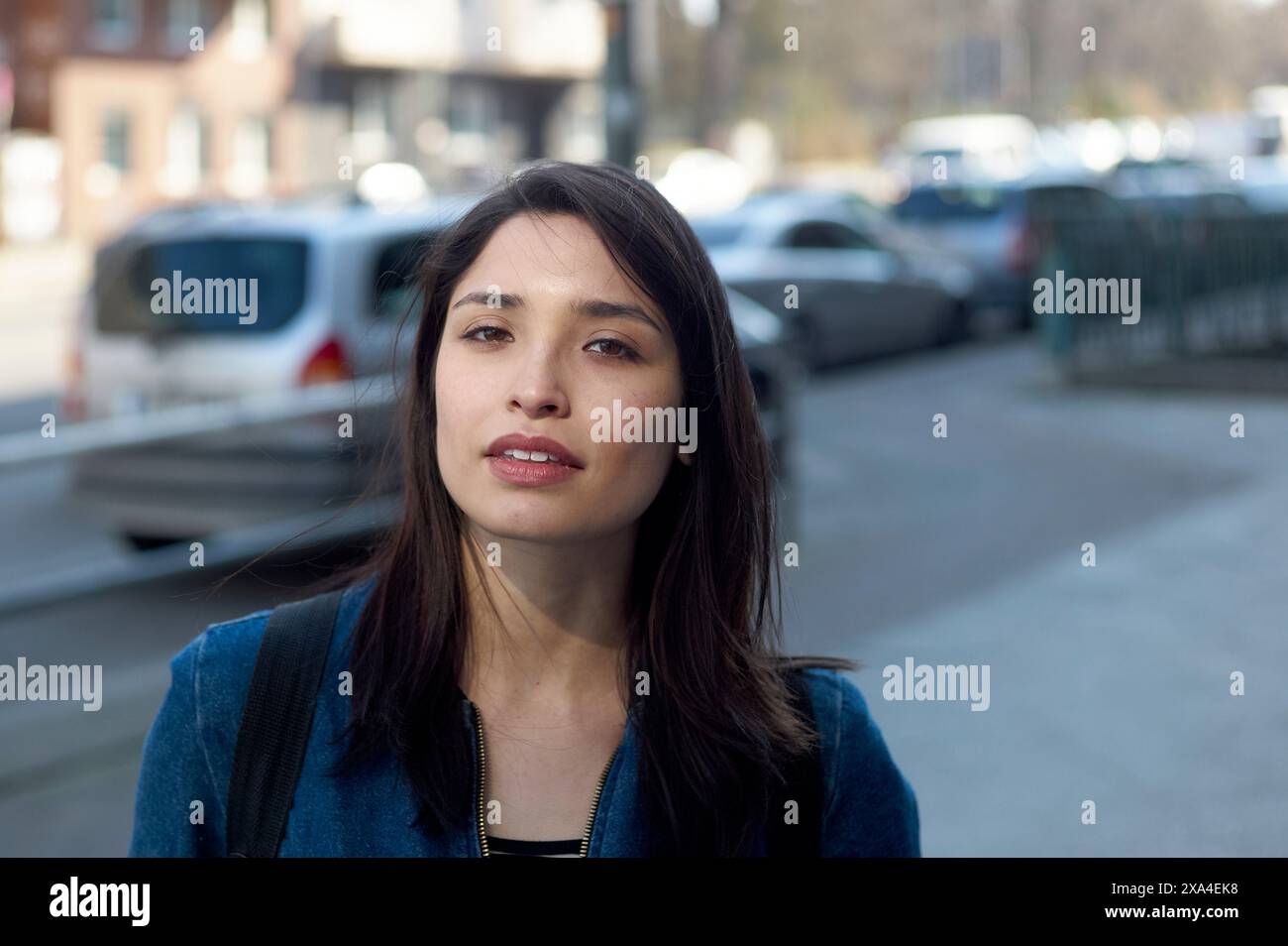 Une femme avec de longs cheveux bruns et une veste en denim se tient dehors avec des véhicules et des bâtiments en arrière-plan, regardant la caméra avec un léger sourire. Banque D'Images
