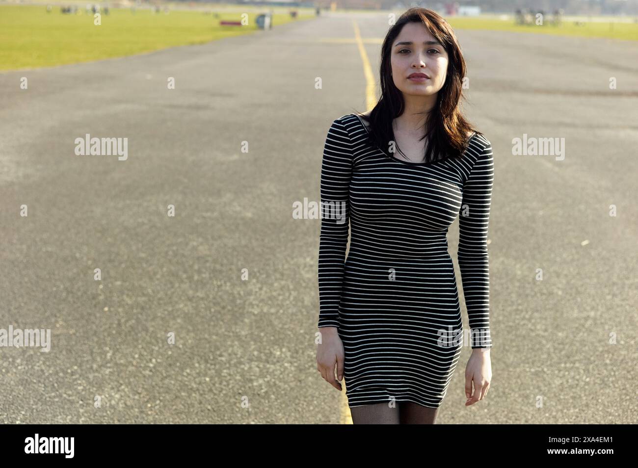 Une femme se tient debout sur une route vide avec des champs d'herbe en arrière-plan, portant une robe rayée noire et blanche. Banque D'Images