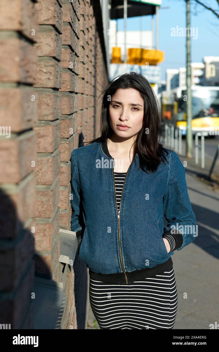Une femme se tient près d'un mur de briques par une journée ensoleillée, portant une veste bleue et une robe rayée, avec une expression contemplative. Banque D'Images