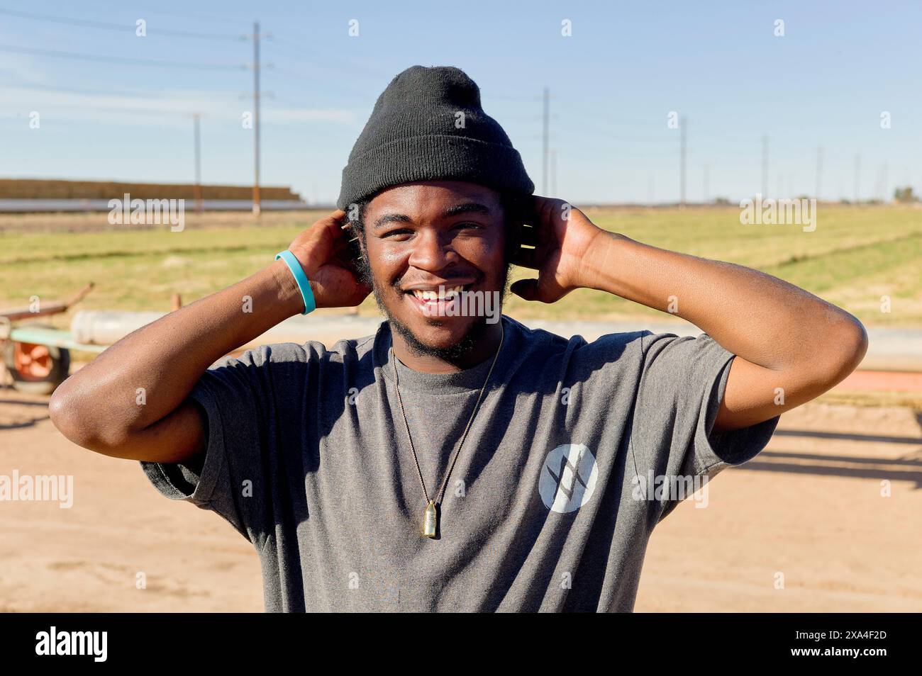 Un jeune homme souriant se tient dehors portant un bonnet noir et un t-shirt, avec un ciel bleu ensoleillé et un champ ouvert en arrière-plan. Banque D'Images