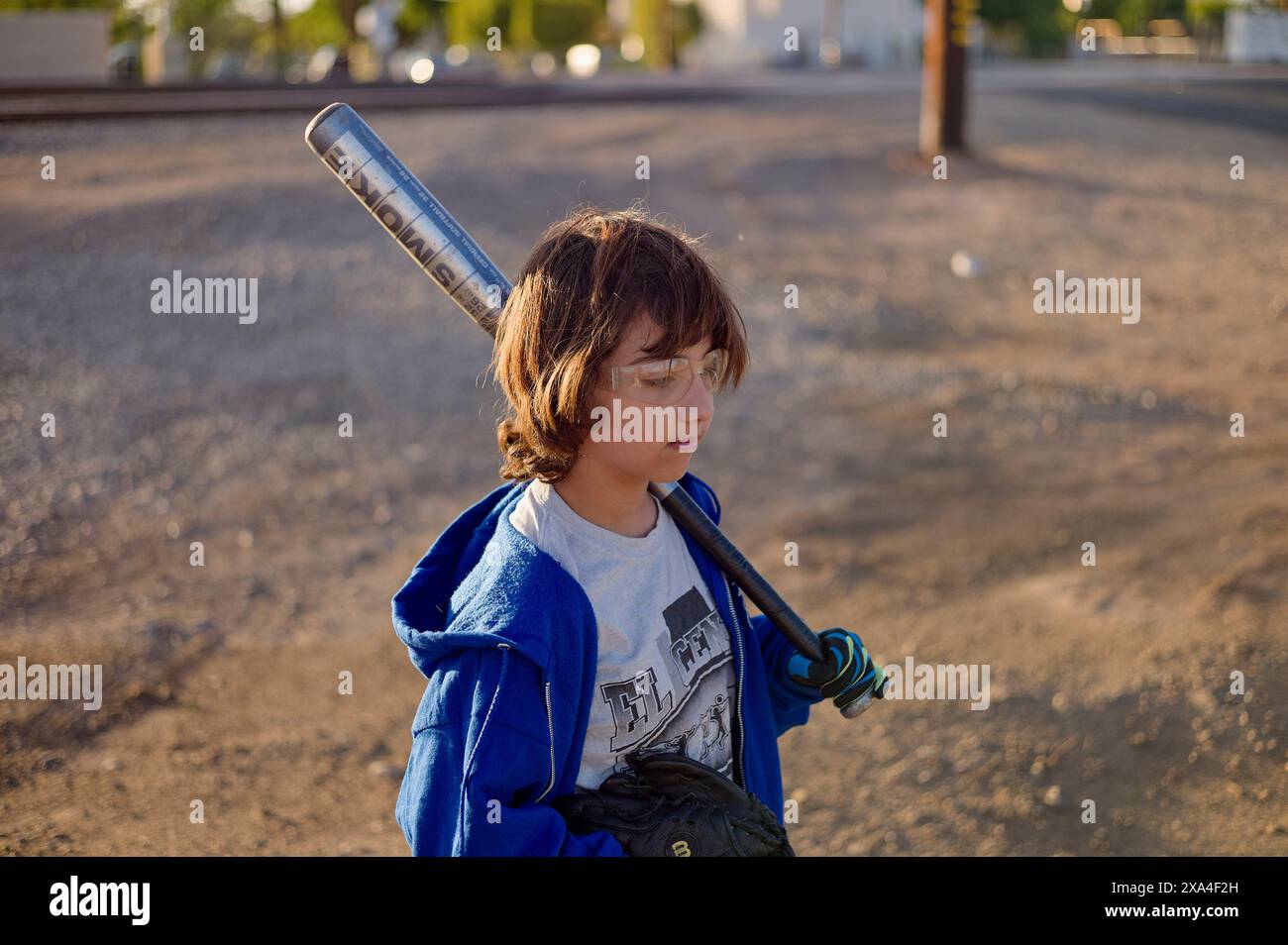 Un enfant avec des lunettes, portant une veste bleue et tenant une batte de baseball sur son épaule, se tient sur une route poussiéreuse au coucher du soleil. Banque D'Images
