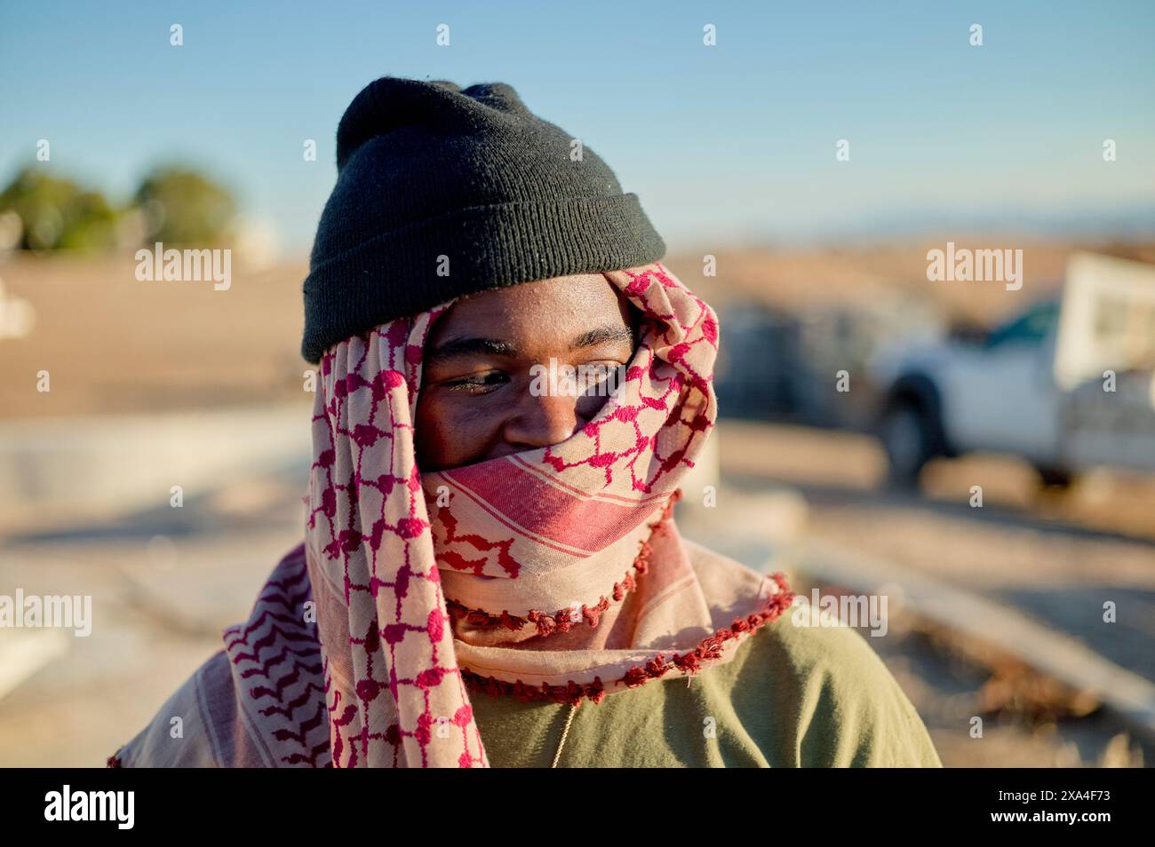 Une personne souriante, enveloppée dans une écharpe à motifs, portant un bonnet, et debout dehors par une journée ensoleillée avec des véhicules et des bâtiments dans le fond flou. Banque D'Images