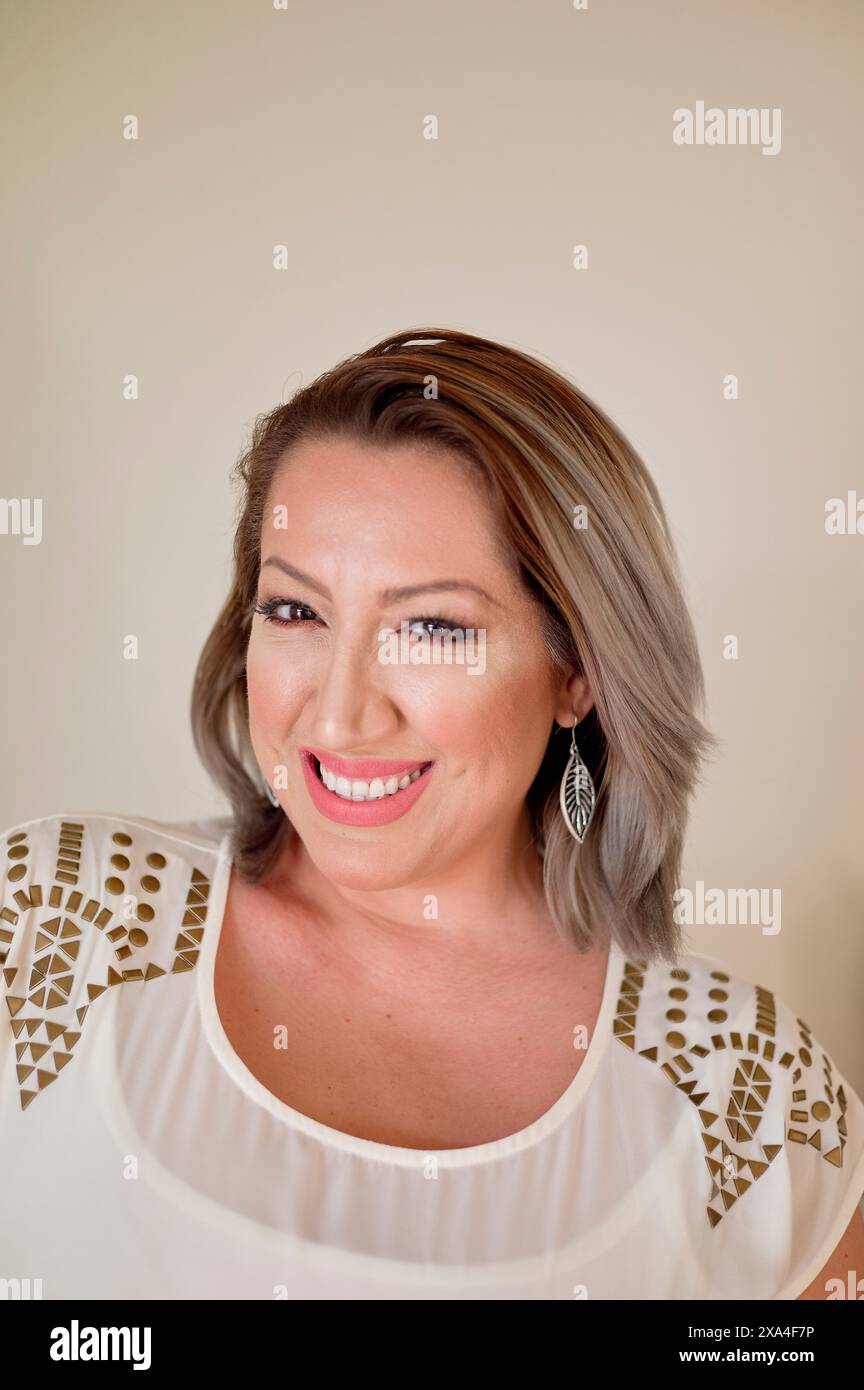 Une femme souriante avec des cheveux gris à la longueur de l'épaule, portant un haut blanc avec un motif d'épaule et des boucles d'oreilles en plumes, pose sur un fond beige clair. Banque D'Images