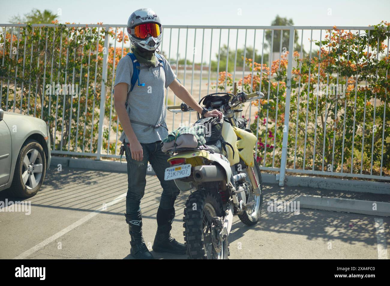 Un motocycliste se tient à côté d'une moto jaune garée par une journée ensoleillée, portant un casque, des gants et un sac à dos. Banque D'Images