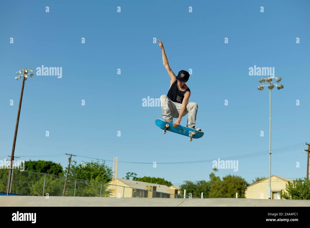 Un skateur effectue un tour en plein air contre un ciel bleu clair dans un skate Park, avec des lumières de terrain de sport et des arbres en arrière-plan. Banque D'Images