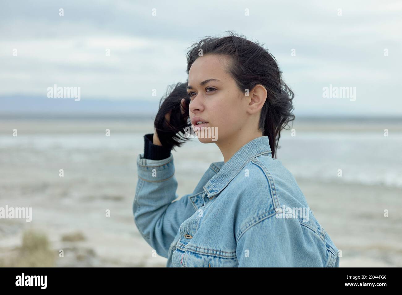 Une femme dans une veste en denim se tient à l'extérieur, avec un plan d'eau serein en arrière-plan, regardant au loin. Banque D'Images