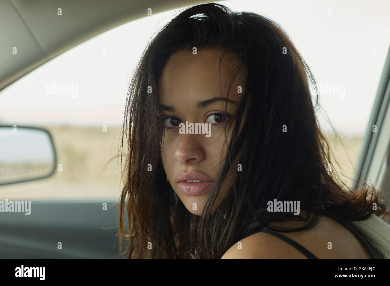 Une femme est assise dans une voiture, se tournant pour regarder la caméra avec un regard intense, ses cheveux foncés émoussés et une expression délibérée et pensive sur son visage. Banque D'Images